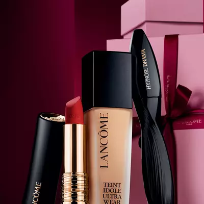 O natură moartă elegantă cu rujul mat L'Absolu Rouge, fondul de ten Teint Idole Ultra Wear și rimelul Hypnose Drama de la Lancome, plasate pe un fundal festiv de lux, de culoare roșu inchis.