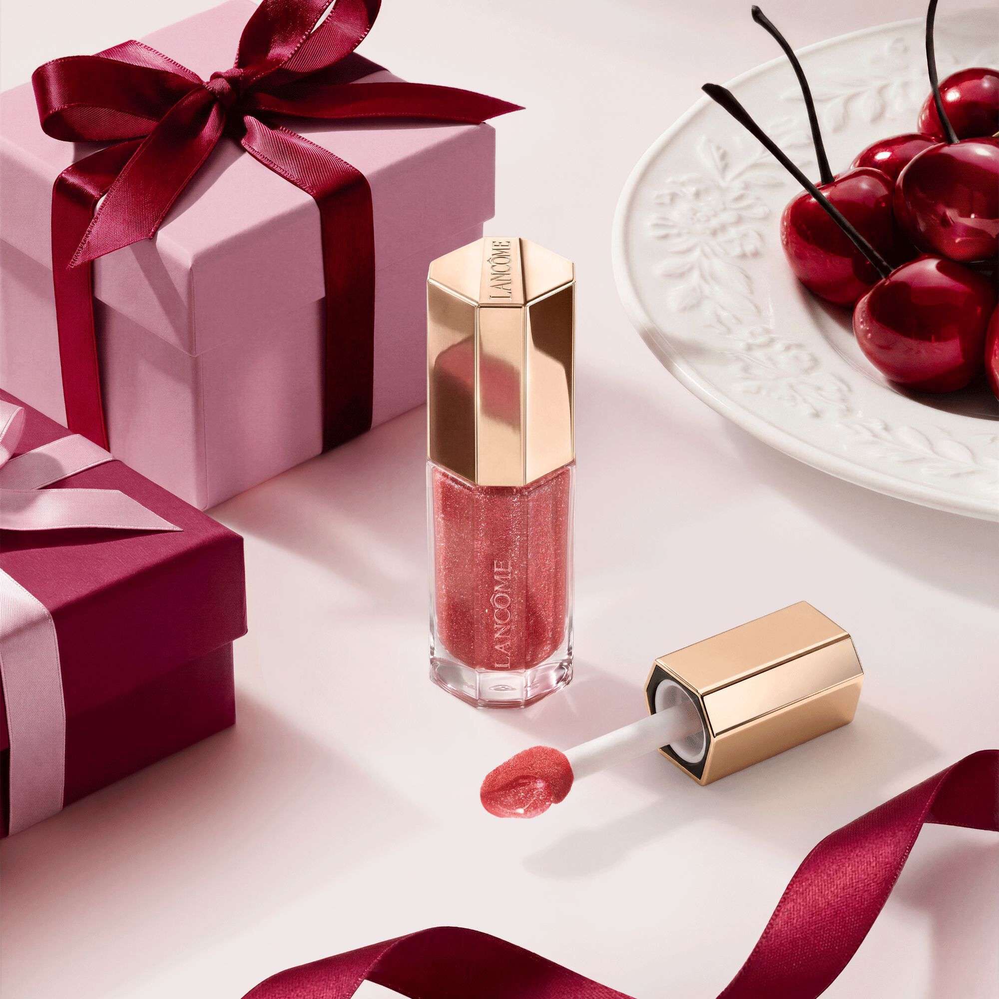 Luciu de buze Lancome in tonuri de fructe de pădure, intr-un tub hexagonal premium, decorat cu cutii cadou și cireșe.