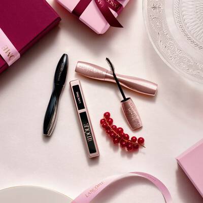 Un aranjament flat-lay cu produse de machiaj Lancome, incluzand mascara și gloss pentru buze, alături de cutii de cadou roz, panglici de satin și o crenguță de coacăze roșii.