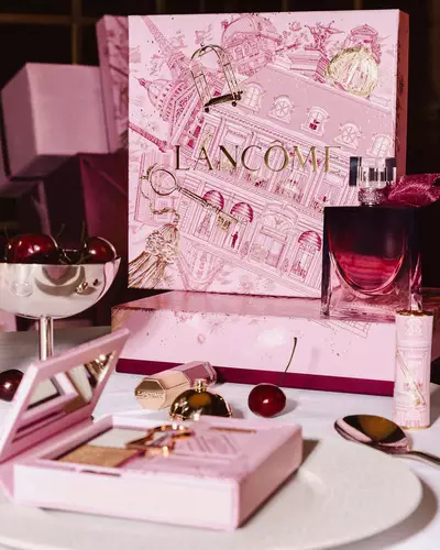 O vitrină festivă de sărbători Lancome, cu o cutie mare de cadou roz ilustrată, parfum și produse de machiaj aranjate pe o masă cu cireșe proaspete.