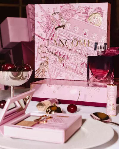 O colecție festivă de sărbători Lancome pe o masă, cu o cutie mare ilustrată roz, parfum, produse de machiaj și cireșe proaspete.