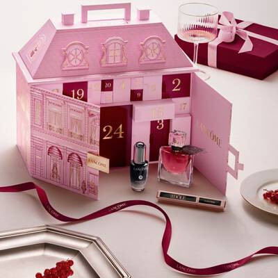 Un calendar advent Lancome in formă de clădire elegantă pariziană deschis, expus alături de produse de infrumusețare și o fundă roșie.