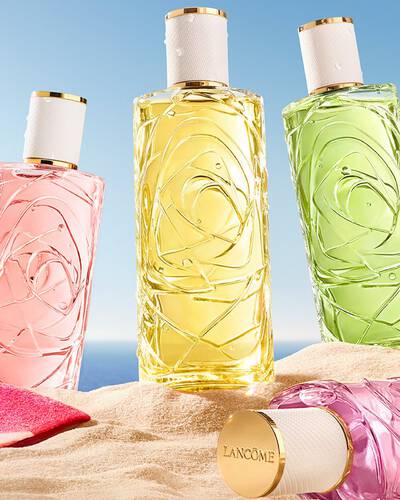 Două flacoane roz - unul din sticlă transparentă cu design texturat, celălalt tip spray - așezate pe o plajă nisipoasă. Un prosop de plajă in dungi roz este așezat alături, sub un cer senin care intalnește linia oceanului la orizont.