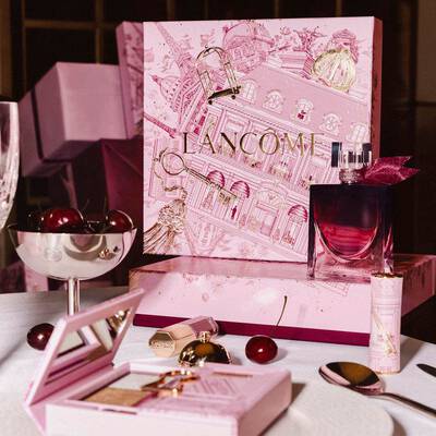 O colecție festivă de sărbători Lancome pe o masă, cu o cutie mare ilustrată roz, parfum, produse de machiaj și cireșe proaspete.