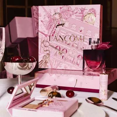 O colecție festivă de sărbători Lancome pe o masă, cu o cutie mare ilustrată roz, parfum, produse de machiaj și cireșe proaspete.