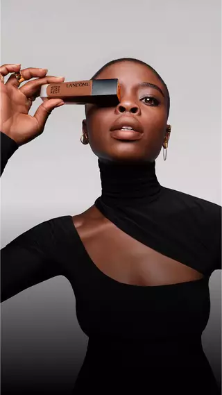 O femeie care poartă un top negru sofisticat, ținand o cutie de makeup Lancome in fața ochiului, cu un zambet elegant pe chip.