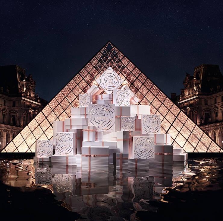 A pyramid of beautifully wrapped gift boxes with rose designs is displayed in front of the illuminated glass pyramid of the Louvre at night, reflecting in the water below.