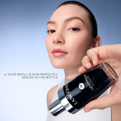 O femeie aplică un ser de față Lancome, ținand un flacon elegant, pe fond alb-albastru.
