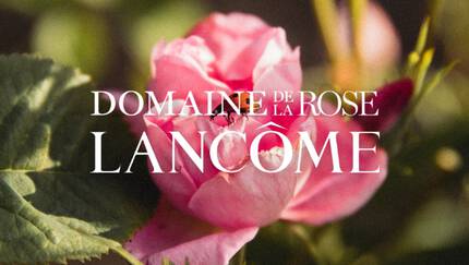 A close-up of a pink rose blossom with green leaves, featuring the text "Domaine de la Rose Lancome" overlayed in elegant font.