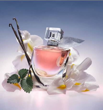 Sticlă de parfum elegantă, de culoare roz, inconjurată de flori albe și frunze verzi, cu un băț de vanilie langă.