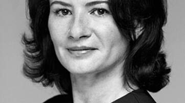 A black and white portrait of a woman with shoulder-length dark hair, wearing a sleeveless dress and looking confidently at the camera.