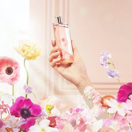 Un flacon de parfum roz este ținut de o mană, inconjurat de flori colorate intr-un decor luminos.