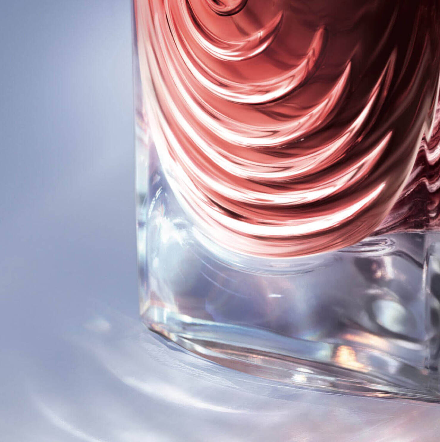 Close-up of a glass bottle with a textured design, featuring rippled layers in shades of pink and clear glass, reflecting light on a smooth surface.