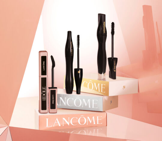 Several Lancome mascara products are displayed on a layered background of soft pastel colors, featuring sleek black packaging and elegant branding.