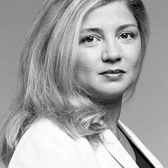 A close-up portrait of a woman with long, wavy hair, dressed in a white blazer, looking confidently towards the camera against a neutral background.