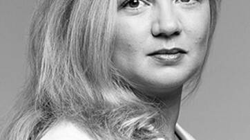 A close-up portrait of a woman with long, wavy hair, dressed in a white blazer, looking confidently towards the camera against a neutral background.