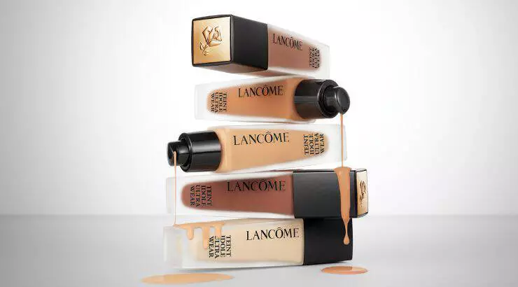 A stack of Lancome foundation bottles in various shades, with some bottles tilted and liquid foundation dripping from them, set against a neutral background.