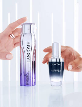A pair of hands holds two skincare products from Lancome: a purple bottle labeled "Renergie" and a silver bottle labeled "Advanced Genifique," both positioned on a reflective surface.