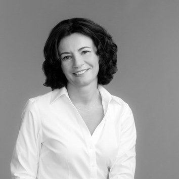 Femeie cu păr ondulat, imbrăcată in cămașă albă, zambind pe fundal gri.