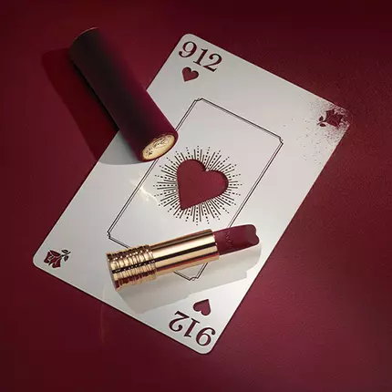 O carte de joc cu un inimă roșie, plasată pe o suprafață burgundă, alături de un ruj roșu inchis.