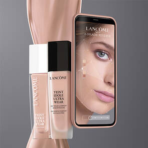 Imagine care promovează un produs de machiaj Lancome, incluzand un flacon de fond de ten și un telefon cu o aplicație care arată un parcurs al feței.