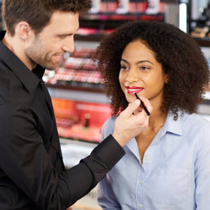 A makeup artist applies red lipstick on a woman sitting in a beauty store, surrounded by various makeup products displayed on shelves.