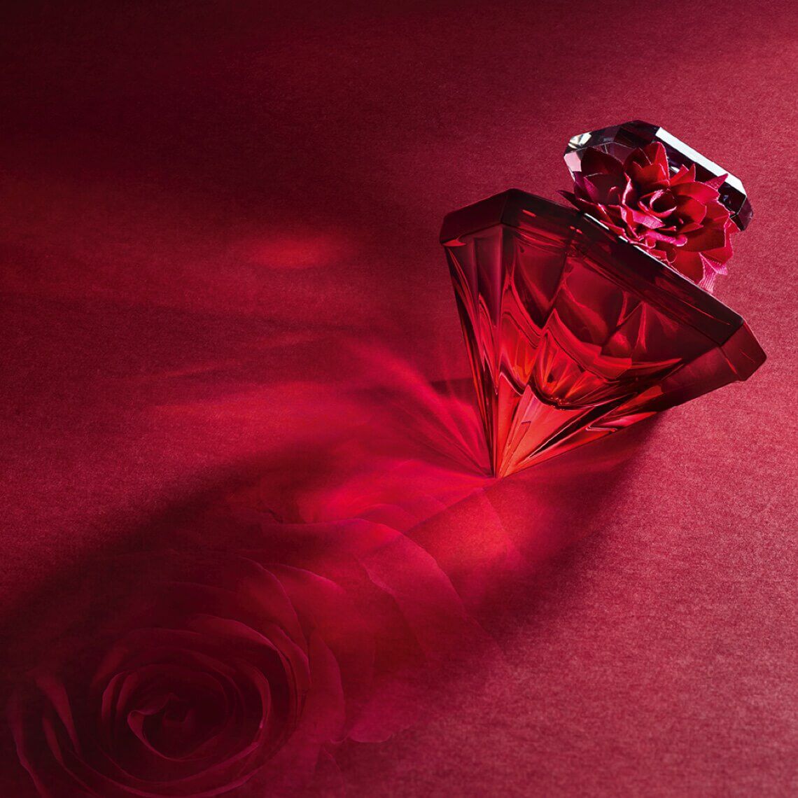 A luxurious red perfume bottle with a diamond-shaped cap casts a soft shadow of a rose on a deep red background.