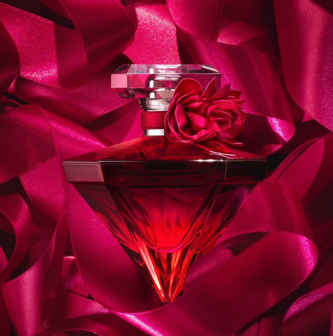 A red glass perfume bottle shaped like a diamond, adorned with a red flower, surrounded by luxurious pink satin ribbons.