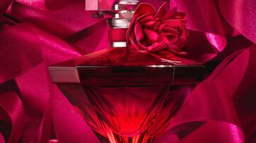 A red glass perfume bottle shaped like a diamond, adorned with a red flower, surrounded by luxurious pink satin ribbons.