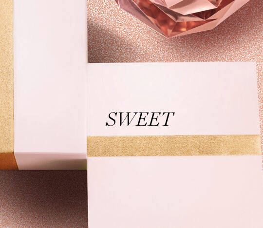 A close-up of elegantly wrapped gift boxes with a gold ribbon, accompanied by a sparkling rose gold decorative item, featuring the word "SWEET" displayed prominently.