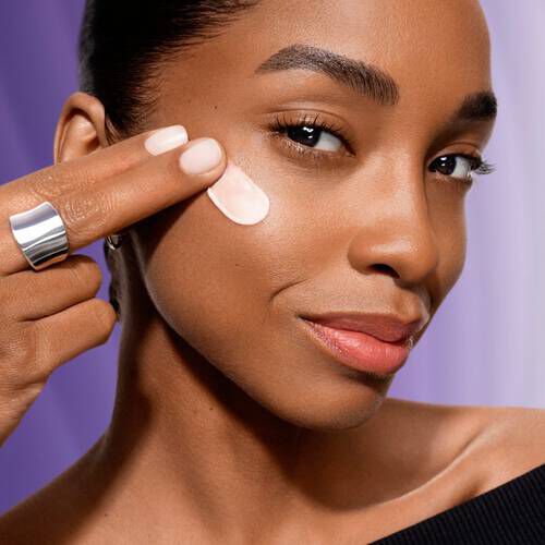 O femeie aplică loțiune pe față, zambind, cu un accent pe ingrijirea pielii. Fundal violet bland.