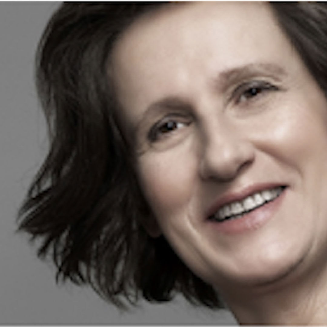 A woman with short, wavy dark hair smiling at the camera against a grey background.