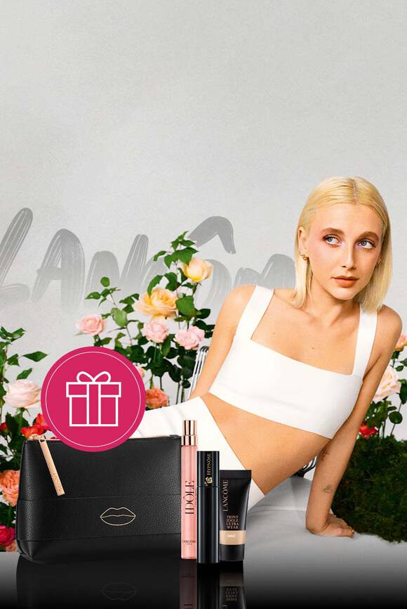 O femeie cu păr blond, imbrăcată intr-o rochie albă, stă langă flori colorate, avand alături o geantă neagră și produse cosmetice.
