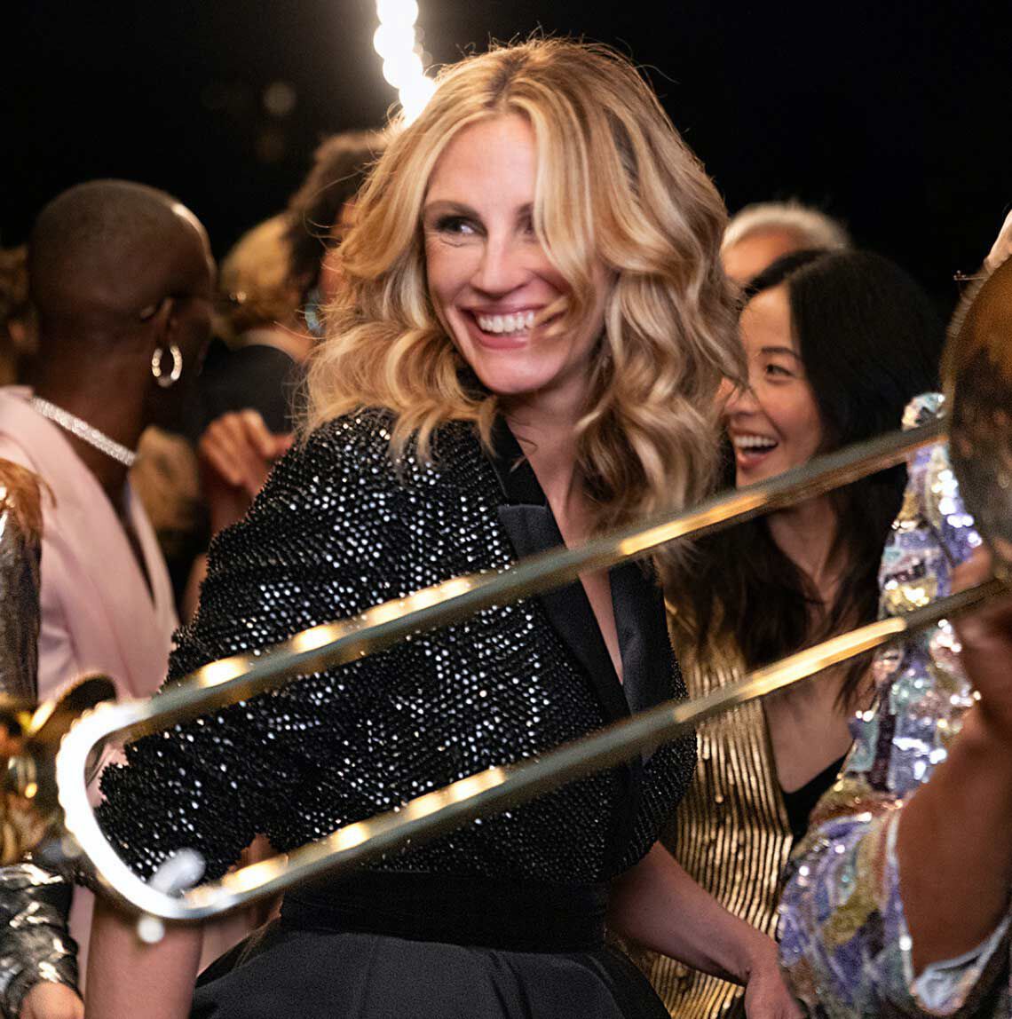 O femeie zambitoare cu părul ondulat, imbrăcată intr-un sacou strălucitor, se bucură de o petrecere alături de alți invitați.