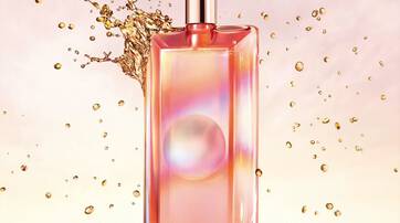 A sleek glass perfume bottle with a pink and gold gradient design, featuring a round logo, surrounded by splashes of golden liquid against a soft pastel background.