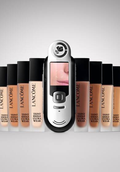 Imaginea prezintă un dispozitiv de testare a nuanței fondului de ten Lancome, plasat in fața mai multor flacoane de fond de ten Lancome, fiecare cu nuanțe diferite.