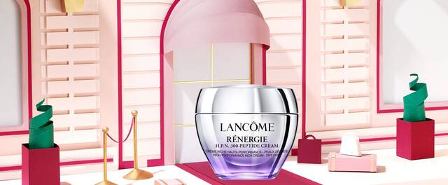 Imaginea prezintă un flacon de cremă de față Lancome Renergie, amplasat pe un suport roșu, intr-un decor elegant cu nuanțe pastelate și forme geometrice.