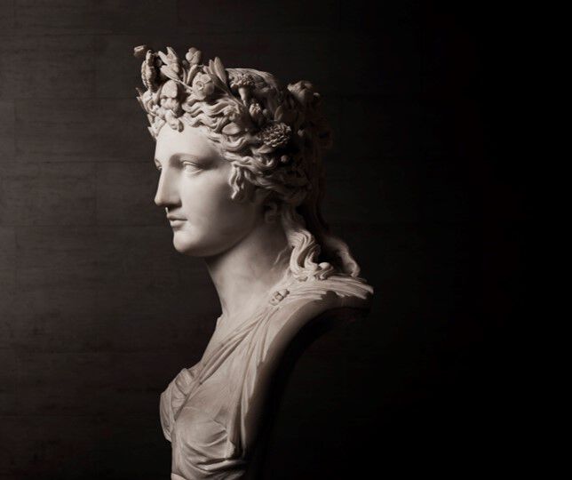 Statuie din marmură reprezentand un bust feminin cu o coroană de flori, evidențiind detalii fine și o expresie calmă, pe un fundal intunecat.