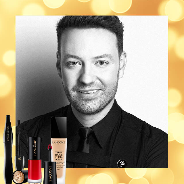 Un stilist de machiaj, zambind, cu produse de cosmetică Lancome aranjate langă el pe un fundal auriu.