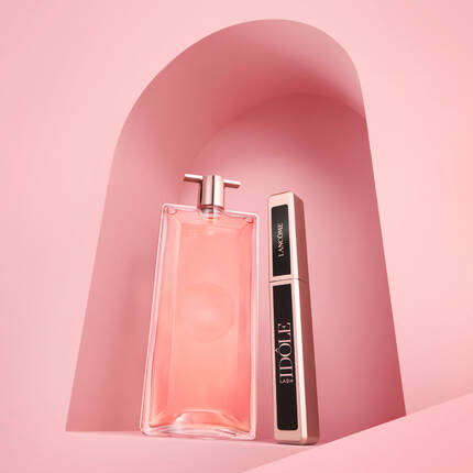 Imaginea prezintă o sticlă de parfum roz și un tus de pleoape, așezate pe un fundal roz cu o formă arcuită.