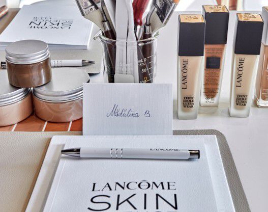 Alt text: Un birou cu produse de machiaj Lancome, inclusiv fonduri de ten și creme, alături de un carnet și un pix alb cu sigla Lancome.