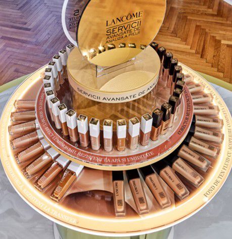 Un stand de produse de machiaj Lancome, cu diverse nuanțe de fond de ten așezate pe un suport rotund, inconjurate de un mesaj despre servicii avansate de analiză a pielii.