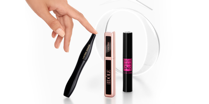 Imaginea prezintă trei tuburi de rimel, incluzand produse de la Lancome, pe un fundal alb, cu o mană care se apropie de un tub negru.