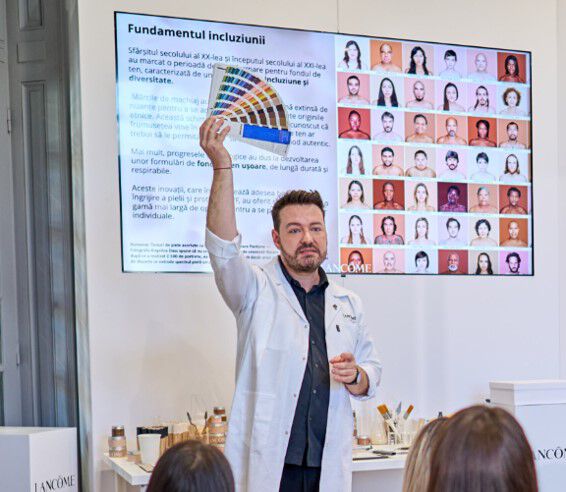 Un prezentator iși ridică un paletar de culori in timp ce vorbește despre incluziune, intr-un eveniment organizat de Lancome, cu un fundal care arată imagini ale participanților.