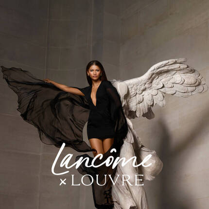 O femeie cu o rochie neagră lungă și aripi albe stă intr-un interior elegant, promovand o colaborare intre Lancome și Louvre.