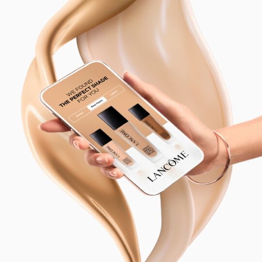 Imaginea prezintă un telefon mobil pe care se află un ecran cu un mesaj despre găsirea nuanței perfecte de fond de ten de la Lancome, inconjurat de un fundal de fond de ten cremos.