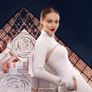 Femeie cu părul blond și machiaj elegant, purtand un pulover alb și buze roșii, pozează langă cutii de cadou și decoruri strălucitoare, cu un fundal al unei clădiri iconică.